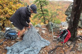 There are hints in this sequence of Kurt Rheinheimer’s brilliant (!)deploying of his rain tarp of what it takes to stop for a great lunch in any season: See silly cap, jillion-dollar rain jacket, waterproof boots and red pack cover still protecting Gail Rheinheimer’s pack contents.